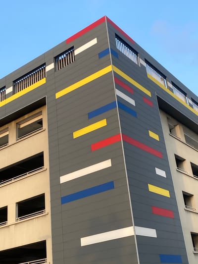 The image captures a low-angle view of a modern building in Falomo, Nigeria. The facade is predominantly gray with vertical stripes of varying widths in red, yellow, white, and blue. The building has several rectangular windows. The sky is a clear, pale blue. The building's design is characterized by clean lines and a minimalist aesthetic.