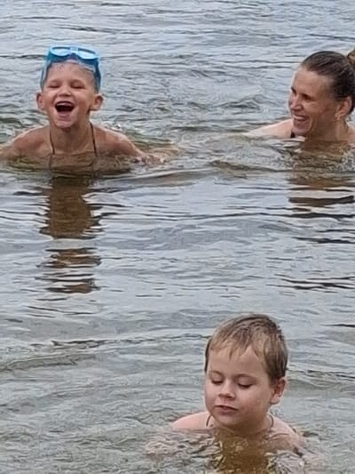 In this media file, three individuals are submerged in what appears to be a body of water, likely a lake or river, given the rippling surface and the natural backdrop of what looks like a wooden structure or shoreline. The scene is oriented vertically.
On the left, a woman with her hair tied up is partially visible, leaning forward and smiling. Her gaze is directed downwards, suggesting she is interacting with someone or something in the water.
In the center-left, a child is in the water, wearing blue swimming goggles. Their mouth is wide open in a joyful or excited expression, and their eyes are closed. The child appears to be having fun.
On the right side of the frame, another child, a boy, is also in the water, his eyes closed and a relaxed expression on his face. His head is tilted slightly, and he seems to be enjoying the water.
The water's surface is textured with light and shadow, creating a dynamic visual effect. The overall impression is one of a casual, outdoor recreational activity, likely during daylight hours given the brightness of the scene. The presence of swimming goggles on one child indicates that swimming or water play is the primary activity. The emotions conveyed are primarily positive, with smiles and expressions of enjoyment. No visible text is present in the image. The location context provided is Reutyntsi, Ukraine, which aligns with a natural, outdoor setting.