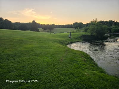 The image depicts a serene sunset scene on a golf course in Bannewitz, Germany. A vast expanse of manicured green grass dominates the foreground, gently sloping towards a body of water on the right side of the frame. The water's surface is partly covered with algae and debris, reflecting the soft hues of the twilight sky. In the middle ground, several trees are scattered across the undulating landscape of the golf course, some forming neat rows along a fairway, while others stand individually. A wooden bench and a small wooden structure with a red seat are visible on the fairway. In the foreground, near the water's edge, a life preserver ring is attached to a post. The sky above is a soft gradient of pale orange and yellow, with a few wispy clouds. The timestamp "19 червня 2026 р. 8:57 PM" is overlaid in the lower-left corner of the image. The overall atmosphere is tranquil and peaceful, characteristic of a late evening in a rural setting.