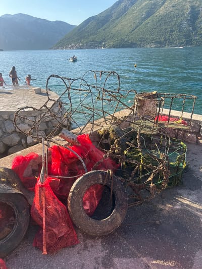The image depicts a coastal scene in Prčanj, Montenegro, under clear skies with the sun low in the sky, casting shadows to the left.
In the immediate foreground, a concrete pier or embankment extends into a body of water. Piled on this concrete are several weathered and corroded items, including multiple fishing or crab traps constructed from rusted metal frames, some covered in marine growth. Interspersed with these traps are several discarded vehicle tires and multiple red mesh bags, some appearing to contain dark, indistinct objects. A corroded metal pipe or rod is also visible among the debris. The concrete surface is discolored and shows various stains.
In the midground, the calm, blue-green water of a bay extends from the pier. Four individuals, appearing to be young females, are present near the water's edge on the left. One person in a pink swimsuit is partially visible entering the water. Another, wearing a black bikini bottom, is stepping into the water. A third, in a patterned swimsuit, wades in the water, facing away from the camera. A fourth person is partially obscured behind the first. Further out in the bay, a small motorboat carrying multiple passengers is moving towards the left, leaving a wake behind it. A single yellow buoy floats in the water to the right of the boat.
The background is dominated by large, steep mountains covered in green vegetation, rising from the water's edge. On the opposite side of the bay, nestled at the base of the mountains, a cluster of buildings forms a distant settlement. The sky above is a clear, light blue.