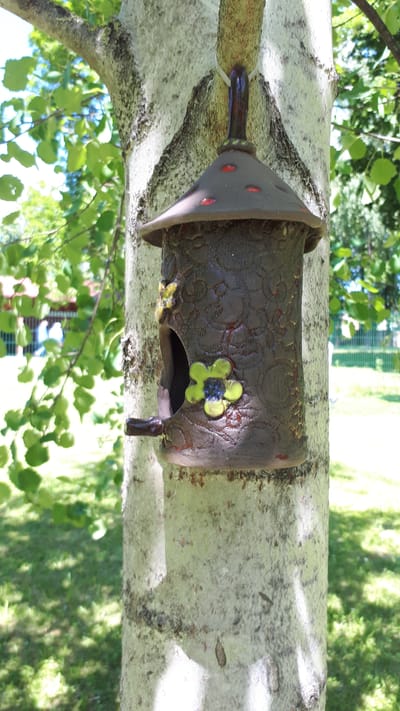 A ceramic birdhouse with a conical roof and a rounded body is attached to the trunk of a light-barked tree. The birdhouse is dark brown with a textured pattern of raised floral designs. Two decorative ceramic flowers, one yellow and one green with a blue center, are affixed to the front of the birdhouse, above and below an oval-shaped opening. A short perch extends from the bottom of the opening. The roof is a darker brown with three red dots. The birdhouse is suspended by a white strap looped around the tree trunk. Sunlight filters through the green leaves of a tree in the background, casting dappled shadows on the grass. In the distance, a blurred fence and building are visible, suggesting an outdoor setting, possibly a park or yard, within Daugavpils, Latvia.