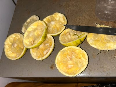 The scene is set in Egbe, Nigeria, on a utilitarian, dark gray or brown textured surface, possibly a countertop or table. Seven halves of squeezed citrus fruits, likely limes or lemons with pale yellow pulp and green-yellow rinds, are scattered across the surface. Visible seeds are present within the disrupted pulp of several halves. A dark-bladed knife, approximately 15-20 cm in length, rests diagonally across two of the fruit halves near the top right quadrant. A clear, ridged plastic bottle is partially visible in the upper right background. No individuals are present in the image.
