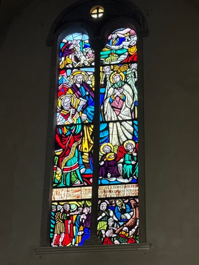 The image depicts a tall, arched stained-glass window within the Duomo di Foligno (Cathedral of San Feliciano) in Foligno, Italy. The window is composed of vibrant, multi-colored glass set within a dark grey stone framework against a plain, light grey interior wall. A small, quatrefoil-shaped window is positioned at the apex of the arch.
The main window is divided vertically into two lancets, each containing multiple horizontal panels depicting various religious scenes with figures wearing halos, indicative of saints or divine beings.
* **Upper Registers:** The topmost panels are less distinct. The upper left shows multiple figures in celestial settings. The upper right depicts figures, one possibly Christ-like, interacting in a heavenly context, with elements suggesting clouds or a divine realm.
* **Middle-Upper Registers:**
* The left lancet features a prominent, bearded figure with a halo, clad in purple and blue robes, holding a staff or scroll-like object. Below him, another bearded figure with a halo, in red and green robes, firmly grasps a large golden key, identifiable as Saint Peter.
* The right lancet displays a tall, slender figure with a halo, dressed in flowing white and pink garments, with hands clasped in prayer or devotion. This figure is flanked by smaller angelic figures with wings.
* **Middle-Lower Registers:** Below the main figures, both lancets feature smaller, seated angelic figures with halos and wings, depicted in purple and green robes.
* **Bottom Registers:**
* The lower left panel portrays a dynamic group scene. A central figure in red and green interacts with multiple individuals, possibly a crowd, in various colored robes. The interaction appears to be a sermon or a miracle.
* The lower right panel shows a more tumultuous scene. A figure in blue and yellow engages with prostrate or struggling individuals, one of whom is entangled with a brown, beast-like creature or demon, suggesting an exorcism, a healing, or a struggle against evil.
* **Inscriptions:** Latin text is visible on horizontal bands beneath the middle-upper registers. The right inscription reads, "DECAN. D. DOMENICO FEDE III PARROCCHIANI DONARONO NEL 25 DI SACERDOZI" (Dean D. Domenico Fede and the parishioners donated on the 25th anniversary of priesthood), confirming the ecclesiastical context and the location in Foligno Cathedral.
The window is backlit, allowing natural light to pass through the colored glass, illuminating the detailed artistry and vibrant hues of the religious narrative.