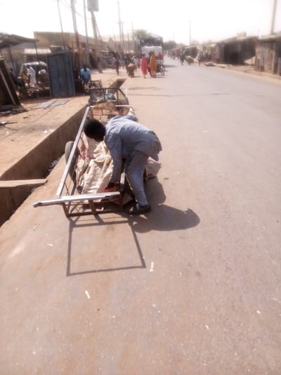 The image depicts a daytime street scene in Tudun Murtala, Nigeria, under bright sunlight with atmospheric haze. In the foreground, a man with dark hair, wearing a light grey long-sleeved tunic and trousers with dark sandals, is bent over a metal-framed two-wheeled pushcart. He is positioned with his back partially to the viewer, facing right, with his hands near the cart's contents. The cart contains multiple light-colored sacks, appearing to be made of fabric or burlap. The cart is situated on a concrete surface next to a concrete drainage channel on the left, with its right side on a light brown paved road.
The paved road extends into the background, flanked on the left by various structures including market stalls and buildings constructed from corrugated metal and masonry. Several individuals are visible in the midground: one person in a blue garment near the left-side structures, and two individuals in red and yellow/cream garments walking away from the viewer on the left side of the road. Another person is pushing a two-wheeled cart further down the street. A large truck, covered with a dark tarp, is visible further down the street on the left. Utility poles with wires line the left side of the street. The background shows the road receding, bordered by similar structures, under a bright, pale sky.