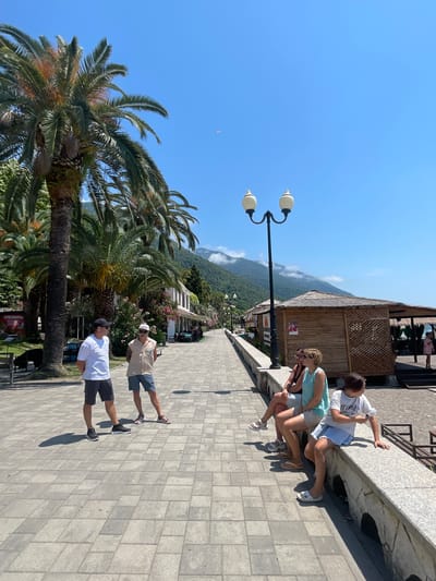 The image depicts a sunny day along a paved promenade in Abaata, Abkhazia. Tall palm trees line the left side of the walkway, with buildings and open-air gazebos visible further down. In the background, lush green mountains rise towards a clear blue sky, with a few clouds obscuring the peaks.
On the promenade, two men in casual attire are standing and talking. One man wears a white t-shirt, dark shorts, and a black baseball cap, while the other is in a sleeveless beige shirt, light shorts, and a white cap. Further along, a group of four people are seated on a low stone wall. Three women are visible: one in a light blue tank top and shorts, another in a black tank top and shorts, and a younger person in a white t-shirt and denim shorts. A fourth person, partially obscured, is also seated. To the right of the promenade, a wooden gazebo with a thatched roof stands on the sand, suggesting it is near the beach. A single street lamp with two lights is prominent on the left side of the walkway. A paraglider is visible in the sky above the mountains.