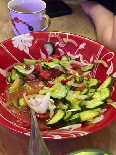 A close-up shot captures a red bowl filled with a fresh cucumber and tomato salad, lightly dressed. Slices of cucumber and tomato are mixed with thin slivers of white onion, all coated in a glistening dressing. The bowl itself features a decorative white floral pattern against its vibrant red background. In the upper left corner, a white ceramic mug with a gold handle sits, adorned with a chain-like design of red and black beads. A portion of a person's leg, clad in dark fabric, is visible in the upper right, suggesting they are seated near the table. A cell phone, with its screen facing down, rests on the table behind the mug. The overall setting appears to be a casual meal, possibly at home. The location is identified as Mineralnye Vody, Russia.