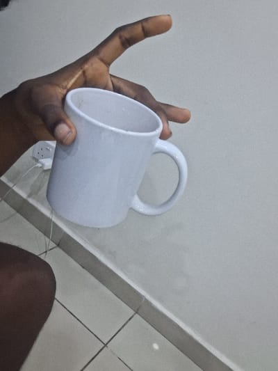 An indoor scene in Close, Nigeria, features a dark-skinned individual's right hand gripping a white ceramic mug. The mug is held at an upward angle, its interior showing discoloration. In the background, a light-colored wall displays a partially visible white electrical wall socket with an inserted white cable. A dark gray baseboard separates the wall from a light-colored tiled floor composed of square sections. The individual's dark-skinned upper leg is partially visible in the lower-left foreground.