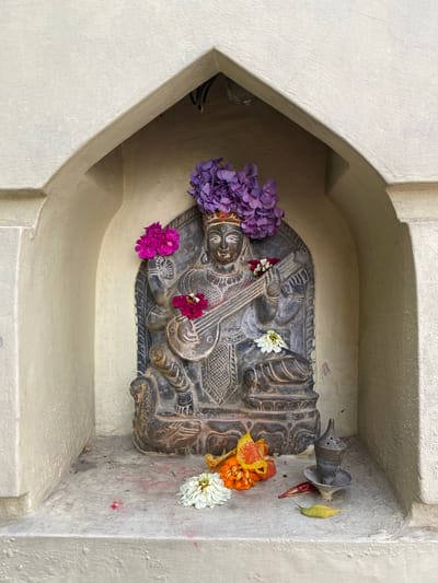 A carved stone statue of a deity, likely Saraswati, is placed in a recessed alcove within an arched, off-white plastered wall. The deity, depicted with multiple arms and holding a stringed instrument, is adorned with purple and pink flowers. The statue is positioned on a ledge that also holds several more flowers, including white and orange blooms, a small metal incense burner, and fallen leaves. The background of the alcove appears to be darker and possibly contains some wiring. The scene suggests a shrine or devotional space. The overall environment is outdoors, with natural light illuminating the scene. The architecture suggests it might be part of a temple complex or a roadside shrine within Kathmandu.