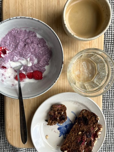 This is a high-angle, close-up, eye-level shot of a breakfast setting on a light-colored wooden cutting board, which is placed on a black and white checkered cloth. The scene features a bowl of purple chia seed pudding topped with yogurt and raspberries, a glass of water, a slice of chocolate banana bread on a plate, and a cup of coffee. The setting appears to be in a home kitchen. The food and drinks are arranged for a meal. No people are visible in the image. The food is the main subject. The lighting is natural and soft. The colors are mostly natural and muted. The overall mood is calm and serene.