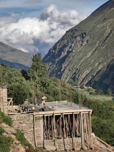 This image depicts a construction site in the mountainous terrain of Tiling, India. The foreground features a partially constructed building with exposed concrete pillars and wooden scaffolding supporting the roof slab. Several workers are actively engaged in construction activities. One worker, wearing a striped shirt and dark pants, stands on a wooden platform, while another, dressed in a light-colored top and dark pants, is positioned near the roof. A third individual, in a green vest, appears to be pouring or spreading concrete. The building is constructed on a stone foundation, blending into the natural landscape.

The background reveals the imposing natural beauty of the region. Lush green trees cover the slopes of steep, rugged mountains, some of which are partially obscured by dramatic clouds. Terraced fields, indicative of agriculture, are visible on the lower slopes of the mountains, suggesting the presence of a village or settlement. The sky is a mix of blue and white clouds, hinting at either a partly cloudy day or the late afternoon light. The overall scene captures a moment of human endeavor against a backdrop of awe-inspiring natural scenery.