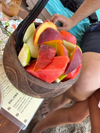 A medium shot from a high angle shows a wooden bowl overflowing with cut fruit, including watermelon, peaches, and possibly apples or pears, placed on a table next to a menu. A black plastic fork is inserted into the fruit. In the background, a person's arm and legs are visible, suggesting they are seated. The environment appears to be outdoors, possibly at a beach or a casual dining setting, given the sandy ground visible at the bottom of the frame. The menu has sections labeled "SALADS" with items like "GREEK SALAD" and "CAESAR SALAD." The overall scene depicts a refreshing fruit snack being enjoyed in a relaxed outdoor setting.