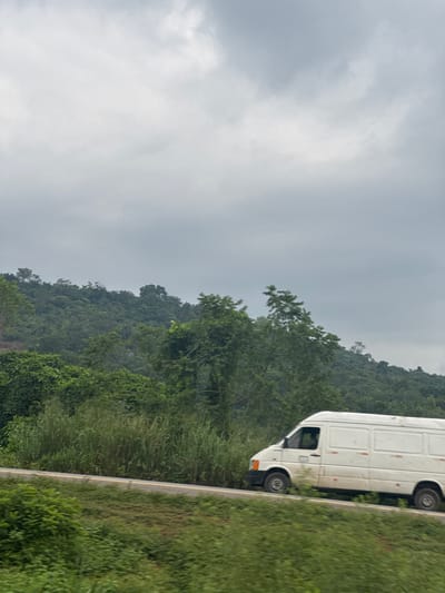 A white panel van is captured in profile, moving from left to right on a paved road. The van appears slightly dirty and has orange and red markings on its sides. The road is bordered by lush green vegetation, including tall grasses and trees. In the background, a dense, tree-covered hillside rises under an overcast, cloudy sky. The image appears to be taken from a moving vehicle, suggesting a roadside perspective within the city of Umuoye, Nigeria.