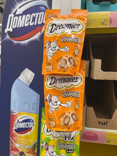 This media file captures a shelf display in a retail setting, likely a supermarket or pet store in Volzhsky, Russia. The primary focus is on various pet food products. Two prominent orange pouches of "Dreamies" cat treats, specifically "Appetizing Chicken" flavor, are displayed side-by-side. These pouches feature cartoon illustrations of cats and chickens, with text indicating they contain "tasty pillows" and high protein content, and are free of artificial flavors. The weight of each pouch is listed as 60g.
To the right of the "Dreamies" pouches, a green package of "Dreamies Mix" cat treats, also featuring "Appetizing Chicken" and "Mint" flavors, is partially visible. Below the cat treats, a large bottle of "Domestos" cleaning product is displayed. The "Domestos" bottle is blue and white and features text in Russian, including "ANTINALET + BLECK" (Anti-Mold + Shine) and mentions grapefruit and pineapple scents. It also states "CHANGES AROMA".
In the background, cardboard retail shelving and packaging are visible, suggesting a typical store environment. A price tag attached to a pink package in the upper left corner shows prices in Russian rubles, with options for "WITH CARD" and "WITHOUT CARD" discounts, further confirming the retail context. The overall scene is brightly lit, typical of indoor retail lighting. No people are visible in the image.