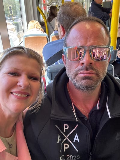 This media file features a selfie of a man and a woman taken inside a public transport vehicle, likely a tram or bus, in Ostend, Belgium, during the daytime under sunny weather. The image is oriented diagonally, with both subjects looking towards the camera.

In the foreground, a woman with blonde hair is smiling broadly, with her face partially visible in the lower right of the frame. She is wearing a pink top or jacket and a delicate necklace.

Beside and slightly behind her is a man with a beard, short hair, and mirrored light blue sunglasses. His sunglasses reflect the vehicle's interior and a street scene, including what appears to be a streetlamp or traffic light. He has a slight smile and is wearing a black zippered hoodie. A thin gold chain is visible around his neck. On the front of his hoodie, there is a prominent white graphic with the word "PAPA" stylized in a cross design, and the text "EST. 2023". A faint "NIKE" logo is also visible on the hoodie's right shoulder.

The background reveals the interior of the vehicle, including yellow handrails, generic seating, and other blurred passengers. Through the windows, an urban or semi-urban environment can be discerned. Both individuals appear relaxed and content, suggesting they are enjoying their journey.