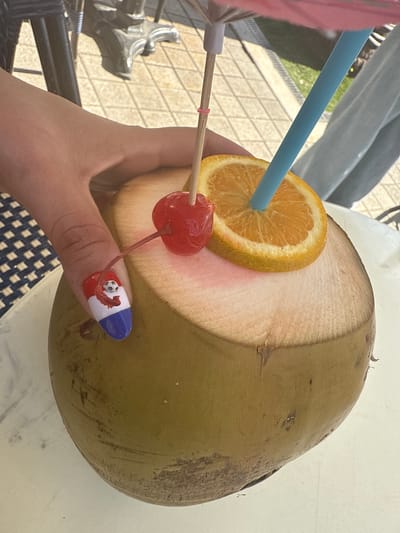 In Miami Beach, United States of America, a person's hand with a red, white, and blue nail design featuring a small soccer ball is holding a fresh coconut. The coconut has been opened and is presented as a drink, garnished with a slice of orange, a maraschino cherry on a cocktail pick, a small pink umbrella, and a blue straw. The scene appears to be outdoors, with a glimpse of a tiled patio and outdoor furniture in the background, suggesting a casual, possibly resort-like setting.