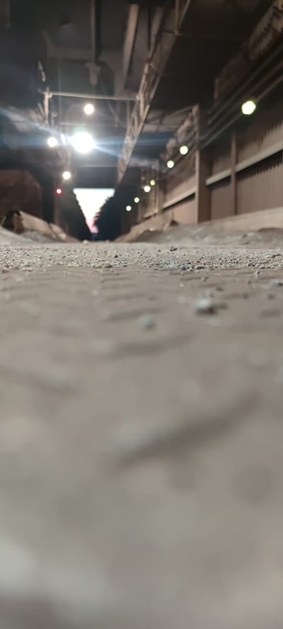 The image depicts a low-angle perspective looking down a gravelly, sandy pathway. The pathway is bordered on the right by a corrugated metal wall and on the left by a sloping earthen embankment. Overhead, an industrial structure is visible, featuring exposed beams and a ceiling with multiple lights. The lights appear to be a mix of bright white and yellowish bulbs, some of which are creating glare. Further down the path, the scene opens up to what appears to be an exit or a wider area with a glimpse of trees and a colorful sky, suggesting either sunrise or sunset. The overall environment suggests an industrial or possibly a mining-related setting within Salzgitter, Germany. The foreground is slightly out of focus, highlighting the gravelly texture of the ground. There are no people or discernible actions taking place in the image.