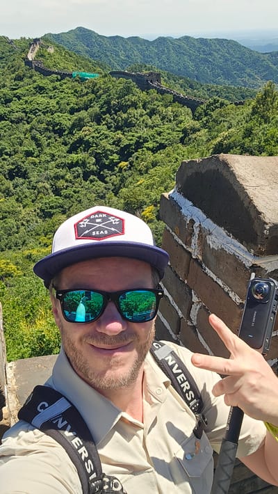 A man wearing a white and blue cap, sunglasses, and a light-colored shirt is taking a selfie on the Great Wall in Huairou District, China. He is smiling and making a peace sign with his right hand. He is holding a black Insta360 X3 camera. The Great Wall stretches across a lush, green, mountainous landscape in the background. The sky is bright and clear.