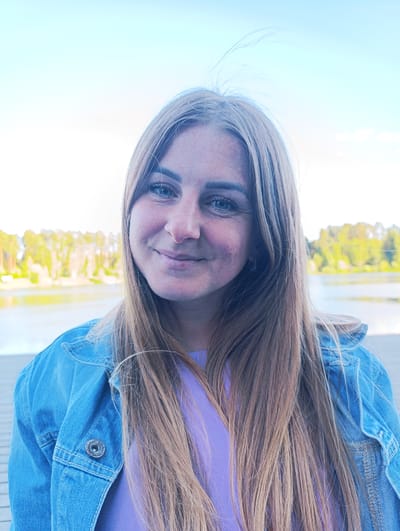 A young woman with long, light brown hair and freckles is captured in a close-up portrait outdoors. She is wearing a blue denim jacket over a purple top and smiling gently at the camera. The background is blurred, showing a body of water and trees under a pale blue sky, suggesting a natural setting within the city of Chernogolovka, Russia.