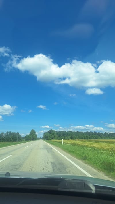 A view from a vehicle moving on a paved road in Glāžšķūnis, Latvia. The road is lined with green grass and trees on both sides, with a yellow field further to the right. The sky is a bright blue with scattered white clouds. There are white lines on the road indicating lanes. The foreground shows the dashboard of the vehicle.