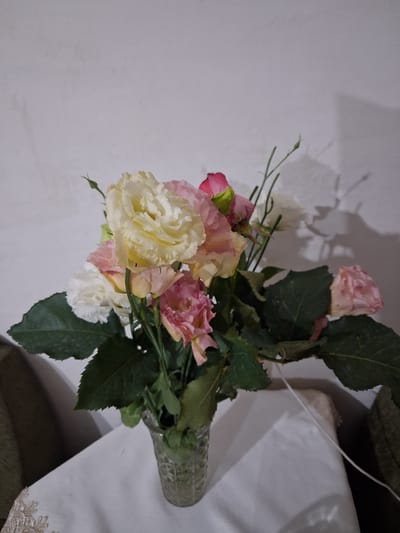 A bouquet of pink and white flowers is arranged in a clear glass vase on a white tablecloth. The flowers are a mix of fully bloomed and budding roses and lisianthus. The vase is placed on a table covered with a white cloth, which has an ornate lace edge on the left side. Behind the vase and flowers, a textured white wall is visible, with shadows cast upon it. The scene is indoors, in what appears to be a domestic setting, likely in the city of Yeghegnadzor, Armenia. No people are present in the image.