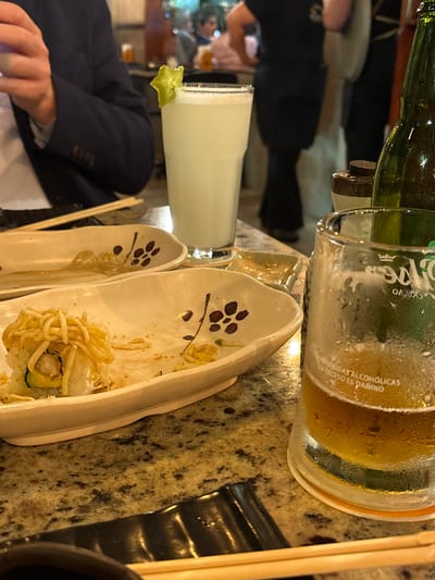 In a restaurant located in Magdalena del Mar, Peru, a table is set with food and drinks. A partially eaten sushi roll sits on a decorative white plate. Another empty white plate with a floral design is beside it. A glass of light-colored beverage with a star-shaped fruit garnish is to the right of the plates. A glass of beer, partially consumed and frosted, is also present. A green bottle of beer stands behind the beer glass. The table surface appears to be granite. In the background, blurred figures of people are visible, suggesting a dining atmosphere. A person wearing a dark blazer is partially visible on the left side of the frame, with their hand on the table.