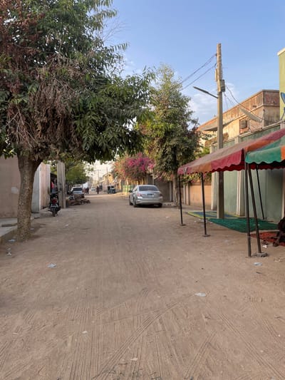 The image depicts a daytime scene on an unpaved street in the city of Daurawa, Nigeria. The street, composed of light brown dirt, shows numerous vehicle tire tracks and scattered debris including paper and plastic.
On the immediate left, a large tree with both green and brown, dried foliage stands, with a black motorcycle parked at its base. Behind the tree, light-colored buildings are visible.
On the right side of the street, a temporary canopy, featuring red, green, and orange fabric, is erected, extending over two green mats and one red mat on the ground. A single individual is partially visible, seated on the ground beneath this canopy, obscured by its support poles. Further along this side, a concrete utility pole equipped with multiple wires and a street light is present. Behind the pole and canopy, a building with a light green lower facade and a textured upper section features a satellite dish and a segment of barbed wire along its top. A light-colored sedan is parked parallel to the curb, facing away from the viewer. Additional trees with green and vibrant pink/purple foliage are interspersed along the right side of the street.
In the distance, the street recedes, revealing several indistinct human figures walking, along with other parked or moving vehicles and additional buildings. The sky above is clear and light blue with minimal cloud cover.