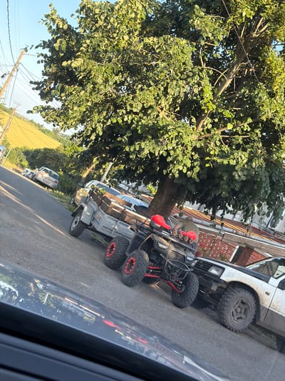 In Tvarditsa, Bulgaria, an outdoor scene depicts a road with parked vehicles and lush foliage. A large tree with dense green leaves dominates the upper portion of the frame, casting shadows on the asphalt. In the foreground, the reflection of the environment can be seen on the glossy surface of a vehicle. A black all-terrain vehicle (ATV) with red accents and large tires is parked on the right side of the road, adjacent to a light-colored utility trailer. Behind the ATV and trailer, a white off-road vehicle with a black hood is parked. Further down the road, several other cars are visible, parked along the side of the street. The background shows a gently sloping hill covered in green fields and trees, under a clear blue sky. Telephone poles and wires are also present, indicating a residential or semi-rural area. The overall impression is one of a quiet, sunlit day in a Bulgarian town.