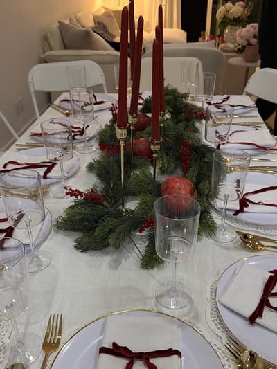A holiday-themed table setting features white plates with gold trim, each adorned with a white napkin tied with a burgundy ribbon. Golden utensils are neatly arranged beside the plates. Clear wine glasses are placed near each setting. A green garland, embellished with red berries and pomegranates, runs down the center of the table, anchoring the arrangement. Burgundy taper candles stand tall within the garland. A floral arrangement in the background adds a touch of elegance to the festive scene.