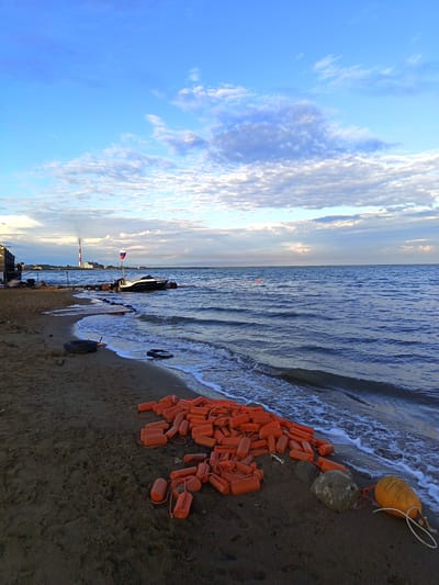 This is a wide-angle, eye-level photograph taken on a sandy beach in Yarovoye, Russia, facing a body of water. The sky is a vibrant blue with scattered white and gray clouds. The water, a deep blue, is gently lapping at the shore, creating white foam.
On the beach, in the foreground and mid-ground, a large pile of bright orange buoys is spread out. Some of these buoys are connected by rope. There are also a few large rocks and what appear to be tire remnants scattered on the sand. The sand itself is dark and appears wet near the water's edge.
In the water, a dark boat with a Russian flag is anchored or tied to a wooden pier. Further in the distance, a tall, red and white striped smokestack is visible, indicating industrial activity. To the left of the smokestack, a low-rise building or structure is present. The overall scene suggests a calm, somewhat industrial coastal environment. There are no people visible in the photograph.