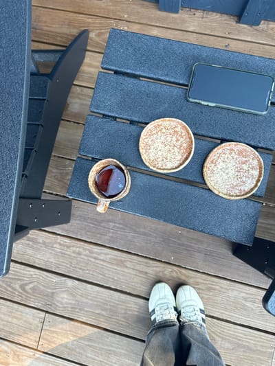 The image depicts an overhead view of an outdoor scene on a wooden deck in Otego, United States. In the foreground, the lower legs and feet of an individual are visible. The individual wears dark gray jeans and white athletic shoes with three black stripes on the lateral sides. Directly in front of the individual, occupying the mid-ground, is a dark gray, slatted outdoor table. On the table, from left to right, are a speckled ceramic mug containing a dark liquid, two matching speckled round ceramic coasters or small dishes, and a light-colored smartphone with a clear case, positioned horizontally with its screen off. Parts of two dark gray slatted chairs are visible on the left and right edges of the frame, flanking the table. The wooden deck flooring shows distinct grain patterns and is illuminated by natural light, casting subtle shadows. No discernible actions or interactions beyond the static arrangement of objects are depicted.