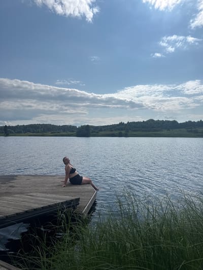 A woman in a black bikini top and black shorts is sitting on a wooden dock, facing the right side of the frame, with her legs extended. She is looking towards the camera with a slight smile. The dock is made of weathered wooden planks and extends into a body of water. The water is calm with ripples and reflects the blue sky. The background features a dense line of trees and vegetation, with a few scattered structures visible. The sky is blue with scattered white clouds. The overall scene suggests a peaceful summer day by a lake or river, possibly in a rural area. The location is identified as Pozdyshki, Russia.