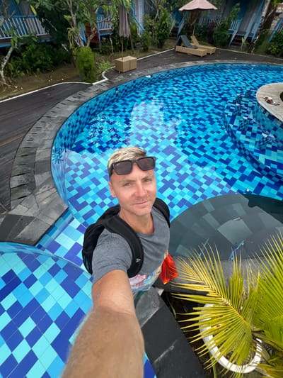 A selfie-style, high-angle shot shows a man with blond hair and sunglasses beside a blue-tiled pool. He wears a grey shirt and a black backpack. To the left are trees, buildings, and lounge chairs. A palm plant in a white pot sits to the right. The pool's design features a pattern of light and dark blue square tiles, creating a mosaic effect.