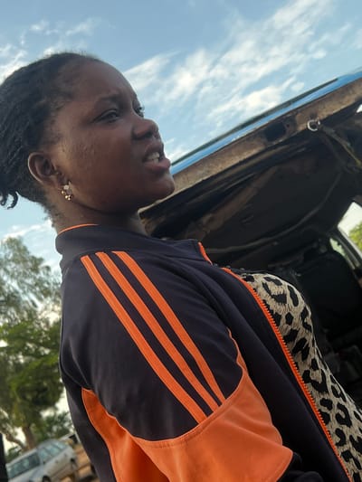 A young Black female is depicted outdoors in Gbaza, Nigeria, positioned with her upper torso and head angled towards the right side of the frame, looking slightly upward. Her mouth is partially open, revealing her teeth. Her dark hair is styled in small braids or twists, pulled back from her face. A gold-toned earring with multiple small, possibly red and white, elements hangs from her visible left ear. Her cheek exhibits some skin texture. She wears a navy blue track jacket featuring three distinct orange stripes running vertically on the left shoulder and sleeve, an orange panel on the lower left front, and a visible orange zipper on the right side. A leopard-print garment is visible beneath the open jacket on her right side. The background consists of a blue sky with scattered white clouds. Immediately behind her is the partially visible, rusted metal frame of the rear section of a vehicle, which has a bluish-green exterior finish and a metal hook attached to its interior rim. In the blurred distant background to the left, green foliage and a light-colored car are discernible.