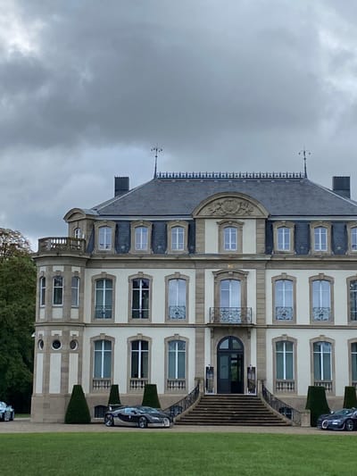 The image presents the Château Saint-Jean in Dorlisheim, France, under an overcast sky. The multi-story building features a light cream-colored facade with light brown stone accents and a dark gray slate roof. The architecture includes multiple rectangular windows with stone surrounds, several dormer windows, and a prominent central entrance accessed by a wide stone staircase. This staircase is flanked by ornate black iron railings. Above the main entrance is a small balcony with a matching iron railing. The roofline is decorated with an intricate black iron railing featuring a fleur-de-lis pattern and two distinct finials. On the left side of the building, a rounded, turret-like extension with a balustraded balcony is visible.

The foreground consists of a manicured green lawn. Two Bugatti supercars are parked on the lawn: a two-tone (silver and dark grey/black) vehicle is positioned to the left of the main staircase, and a dark-colored vehicle is partially visible to the right. Several conical green topiaries are strategically placed near the building and the base of the steps. Green trees are visible in the background on the left side of the scene. No individuals are depicted.