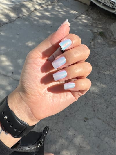 A person's hand with long, square-shaped fingernails painted in a shimmering, iridescent polish is held up in a bright, sunny outdoor setting. The nails reflect shades of light blue, pink, and white, creating a pearlescent effect. The hand has a light skin tone with visible texture and fine lines. A black smartwatch with a silver buckle is worn on the wrist. Partially visible to the left of the hand is a portion of a car's license plate with the text "AU 11". In the background, the ground appears to be a concrete surface with cracks and patches of shadow cast by overhead foliage, suggesting it is daytime with dappled sunlight. The overall impression is a close-up shot focused on the well-maintained fingernails and accessories, likely captured in a casual outdoor environment.