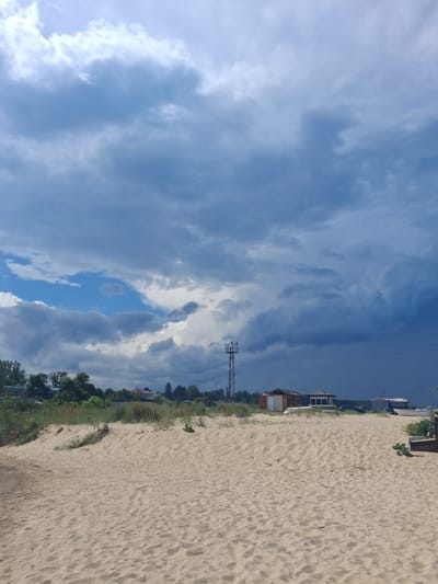 This media file depicts a sandy beach in Shkorpilovtsi, Bulgaria. The scene is dominated by a vast sky filled with dramatic, dark clouds, suggesting a stormy or overcast day, though patches of blue sky are visible.
The foreground is a textured expanse of sand, with some sparse dry grass and twigs scattered across it, indicating it's a natural beach rather than a manicured one. In the middle ground, a line of green vegetation, including trees and taller grass, separates the beach from a few buildings. These structures appear to be simple, low-rise buildings, one with a red tiled roof and another with white siding, suggesting a small settlement or resort area. A prominent metallic lattice tower, possibly a radio or communication tower, stands out against the sky and trees.
The overall impression is one of a slightly wild, natural coastal landscape with the presence of human infrastructure. The time of day is difficult to pinpoint precisely due to the cloud cover, but the lighting suggests it could be late afternoon or early evening. There are no people visible in the image, and no text is discernible. The combination of stormy clouds and the beach setting creates a somewhat somber or dramatic atmosphere.