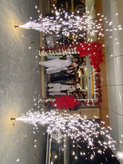 The image shows an event occurring in front of a building, likely in قندهار, Afghanistan, based on the style of dress and the local script on the sign. The entrance to the building is decorated with red floral arrangements forming an arch. A group of men wearing traditional Afghan clothing, including white shalwar kameez and some with head coverings, are gathered at the entrance. Two pyrotechnic devices are in operation on either side of the entrance, emitting streams of sparks. A car is partially visible in the foreground. The setting appears to be a commercial or public space.