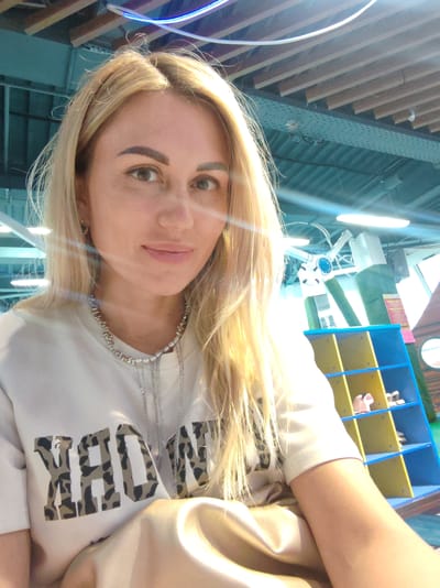The image shows a close-up selfie of a woman with blonde hair and light-colored eyes. She is wearing a white t-shirt with large, leopard-print letters that appear to spell "YORK". She is also wearing a silver necklace with various charms and pendants. The background is blurred but appears to be an indoor environment with wooden ceiling beams and some blue and yellow shelving units with children's shoes visible on them. There is also a clock visible in the background. The lighting suggests it might be daytime with bright artificial lights present. There is no visible indication of the specific location within the city of Amursk, Russia.