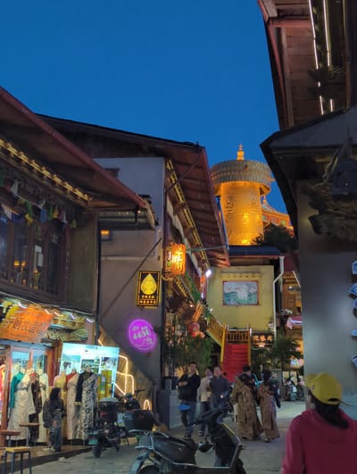 This media file depicts a nighttime scene in what appears to be a street market or shopping area in Shangri-La, China. The image is oriented vertically.

The primary subject is a large, ornate, golden cylindrical structure, possibly a prayer wheel, illuminated by artificial lights, dominating the upper left portion of the frame. Below and to the right of this structure, several buildings with traditional architecture are visible, adorned with neon signs and string lights. One building features a large, colorful painting or tapestry on its exterior.

Several people are present in the scene, some standing and others seated. In the foreground, on the right, a person wearing a yellow cap and a red jacket is partially visible, looking towards the scene. Further back, a group of individuals dressed in what appear to be traditional robes are walking or standing. More people are seen in the background, near stalls and shops. Various objects, including motorcycles, bicycles, and market stalls displaying textiles and goods, are also present.

The activity appears to be a lively evening in a commercial area, likely a tourist spot, with shops open and people browsing. The time of day is clearly evening or night, as indicated by the dark blue sky and the extensive use of artificial lighting. The weather seems to be clear, given the visibility.

Notable details include the intricate design of the golden structure, the vibrant neon signs, and the variety of textiles displayed in the market stalls. The overall mood seems energetic and bustling.

Visible text includes Chinese characters on several neon signs and banners. One sign above a shop reads "普洱茶庄" (Pu'er Tea House). Another sign, in yellow, features a portrait of a person. A purple neon sign displays "440". A banner in the lower right reads "扎染坊" (Tie-dye Workshop).
