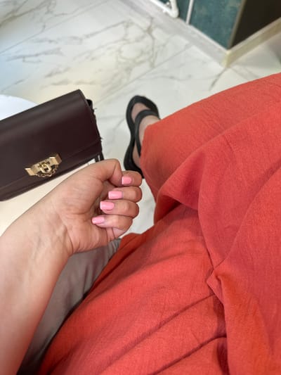 The image shows a close-up view of a person's hand with pale skin and short, light pink painted nails. The hand is resting on a light-colored fabric, likely part of clothing or a cushion. To the left, a dark brown leather purse with a gold clasp is visible. In the background, the person's legs are covered in a bright coral-colored fabric, possibly trousers or a skirt, and they are wearing black flat sandals. The setting appears to be indoors, with a marble-patterned floor and a hint of a green wall in the upper right corner. There is no specific indication of the city of Soroca, Moldova, in the image.
