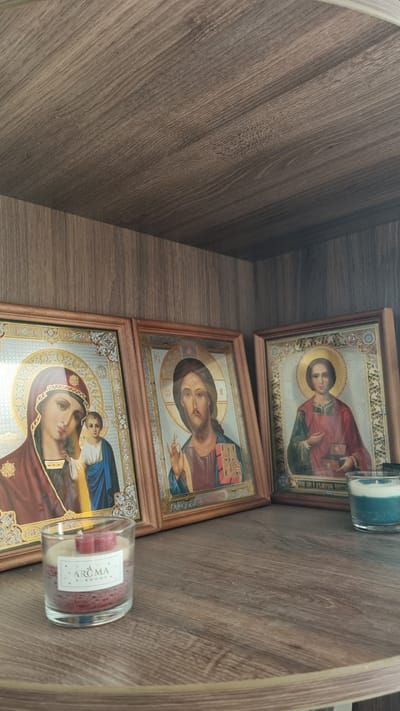 The image shows three framed religious icons and a candle on a wooden shelf. The icons depict the Virgin Mary with the infant Jesus, Jesus Christ, and another male figure, possibly a saint. All figures are rendered in a traditional iconographic style. The shelf is made of a wood-like material with a visible grain pattern. A glass candle with a red and white wax filling and a label that reads "AROMA HARMONY" is placed in the foreground to the left. To the right, a small blue and white candle is visible. The background consists of a darker wooden surface, suggesting this is inside a cupboard or on a bookshelf. The overall setting appears to be domestic. The location is stated to be Верзилово, Russia.
