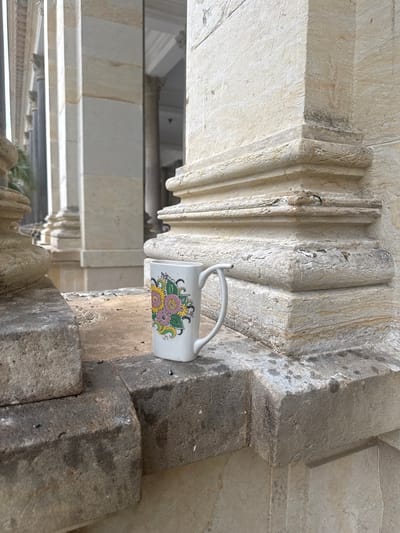 A white mug with a floral design is placed on a stone ledge. The mug is positioned in the foreground, to the right of center. Behind the mug and ledge, a colonnade of classical stone columns stretches into the background, some of which appear to be wet, suggesting a recent rainfall or proximity to water. The architectural style is reminiscent of a grand public building or spa, consistent with the aesthetic found in Karlovy Vary, Czechia. The stone of the ledge and columns is weathered and shows signs of age and exposure to the elements. No people are visible in the image. The lighting suggests it is daytime, possibly overcast or shaded.