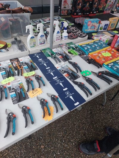 The scene depicts a market stall in Eckbolsheim, France, exhibiting various merchandise. The primary subject is a white table laden with goods. In the foreground and mid-ground, multiple pairs of black and blue/teal handled gardening secateurs/pruners are displayed, both packaged and unpackaged, with several bearing yellow price tags indicating "10€." A row of blue parking discs, each printed with a white "P" and "HEURE D'ARRIVEE," is laid out centrally. To the left, several white plastic spray bottles, labeled as household cleaning products, are visible.
Further back on the table, rows of packaged items, including boxes of Gillette razor blades and small boxes for children's items (e.g., "Goutte d'eau"), are arranged. In the background, additional merchandise is visible, including boxed sets resembling construction toys. A vertical metal pole, likely a support for a market canopy, is present on the table's left side. The ground beneath the table is composed of gravel or small stones. A person's right foot, encased in a dark shoe with a red accent, and part of their dark trouser leg, is visible in the lower right corner, positioned on the gravel next to the table.