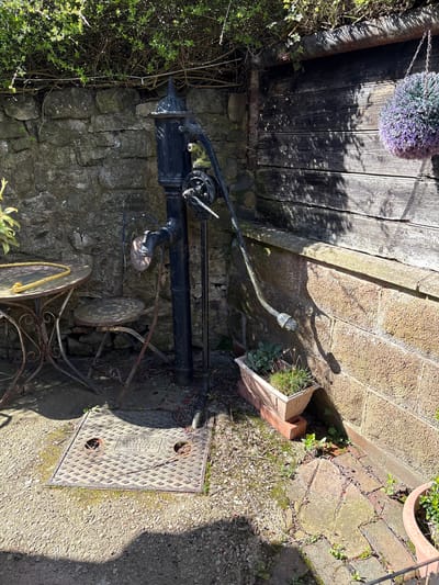 The image depicts an outdoor scene in Shrewsbury, United Kingdom. A black metal water pump stands next to a stone wall and beneath wooden planks. A small, ornate, metal table and chairs are placed near the pump. A metal service cover is on the ground, next to the pump. Vegetation grows near the wall and on the steps. A hanging purple floral decoration is visible above the wooden planks. A brick planter is near the steps. A plant pot is in the top right.