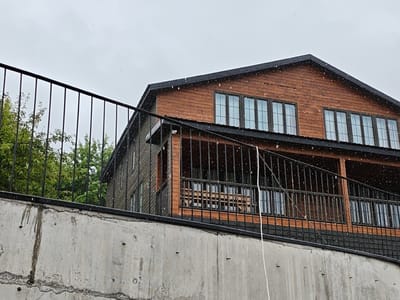 An exterior, eye-level, medium shot captures a two-story wooden house with a dark roof and multiple windows against a cloudy sky. A black metal railing runs horizontally across the frame in front of the house, and a concrete wall is visible in the foreground. The scene is set in Ukeshi, Georgia.