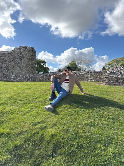 An outdoor, portrait-oriented image depicts a man sitting casually on a vibrant green lawn, leaning against a weathered stone wall. The man, in the foreground and slightly to the left, is looking towards the camera with a slight smile. He is dressed in a light brown jacket, a white sweater with horizontal red and dark stripes, blue jeans, and dark shoes. He also wears sunglasses and a dark beanie hat, and has a beard. His left arm is extended behind him, resting on the grass, while his right arm is bent.
Behind the man, a tall, rough-textured stone wall stretches across the frame, suggesting ancient ruins or a historic structure. Beyond the wall, a few trees with sparse or bare branches are visible, implying late winter or early spring. The upper portion of the image is dominated by a clear, bright blue sky dotted with large, white cumulus clouds. The sunlight is strong, casting a prominent shadow of the man on the grass to his right, indicating either morning or late afternoon. The grass itself is thick and green. There is no visible text in the image. The scene is consistent with the historical landscapes found in King's Lynn and West Norfolk, United Kingdom.