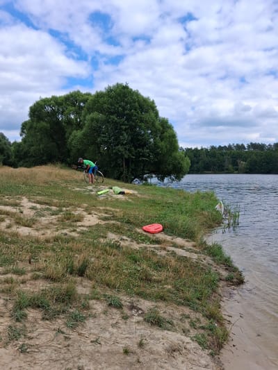 The image depicts a person with a bicycle on the sandy shore of a lake in Reutyntsi, Ukraine. The person, wearing a green shirt and blue shorts, is bent over the bicycle, appearing to adjust it. Nearby, on the grassy bank, are several items including a bright red float, a green cylindrical object, and a lighter-colored bag. The lake's water is rippling, reflecting the cloudy sky, and the opposite shore is lined with dense green trees. The foreground is characterized by dry grass and sand, with sparse vegetation. The overall scene suggests an outdoor recreational setting.