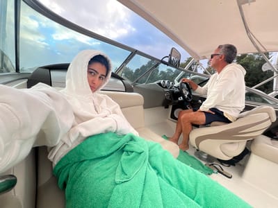 An interior view of a boat in Vila Nova de Cerveira, Portugal, captures two individuals under a partly cloudy sky. In the foreground on the left, a young adult female with dark hair is seated, wearing a white hooded sweatshirt with the hood up, and wrapped in a bright green towel from the waist down. Her right arm is extended, holding the device capturing the image, and she looks towards the viewer with a neutral expression. To her right, an adult male with grey hair and sunglasses is seated at the boat's helm, gripping the steering wheel with his right hand. He wears a white long-sleeved shirt and dark shorts, and is barefoot. His gaze is directed away from the viewer, towards the front of the boat. The boat's interior is white and beige, featuring a windshield, a dashboard with multiple gauges, and a white canopy overhead. A pair of dark flip-flops are visible on the deck near the male's feet. The background shows a shoreline with dense green trees under an overcast sky with some blue patches.