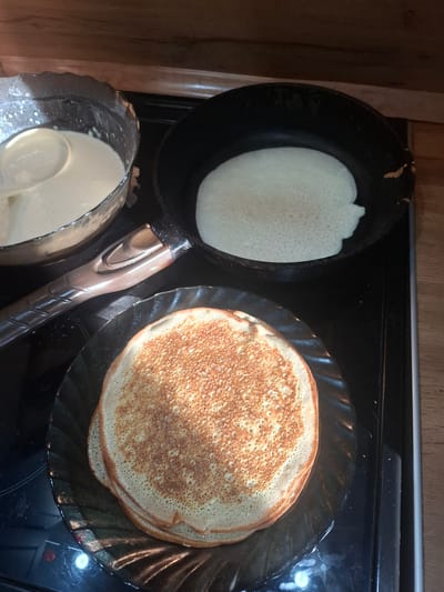 In this overhead shot, a cooking process unfolds on a black electric stovetop, likely in a home kitchen, given the wooden cabinet in the background. The scene is focused on the preparation of what appear to be crepes or thin pancakes.

On the left, a black frying pan holds a pale, bubbly batter, indicating it's in the initial stages of cooking. To the right, a stack of golden-brown pancakes rests on a dark, wavy-edged plate. The pancakes exhibit the characteristic small holes and slightly uneven texture of well-cooked crepes. Below the frying pan, a translucent bowl contains more of the white batter, with a white ladle submerged within it, ready for use. The handle of the frying pan, a textured dark brown and black, is visible between the frying pan and the plate of pancakes.

The lighting suggests an indoor setting, possibly during the day, though no specific time is evident. The overall atmosphere is one of domestic activity, centered around food preparation. There is no visible text in the image. The context of Svobodny, Russia, suggests this culinary scene is taking place in a Russian home.