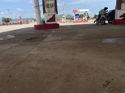 This image depicts an outdoor scene at a gas station in Pasere, Nigeria. The primary subjects are two people on a pink motorcycle parked next to a black car, which is positioned near a gas pump. One person, wearing a green and black checkered shirt, is seated on the motorcycle, while the other person stands behind them. The gas station features red and white structures, including gas pumps with visible "Shafa" branding. In the background, there are hills and a clear blue sky with scattered clouds, suggesting it is daytime with fair weather. Utility poles and vegetation are also visible, contributing to the setting. The ground is paved concrete, showing some wear and tear. A white and red plastic bag lies on the ground in the foreground. The overall impression is a typical scene at a gas station.